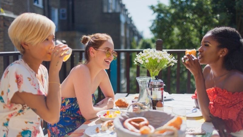 Três amigos desfrutam de uma tarde de Brunch em um terraço enquanto bebem refrigerantes e comem aperitivos